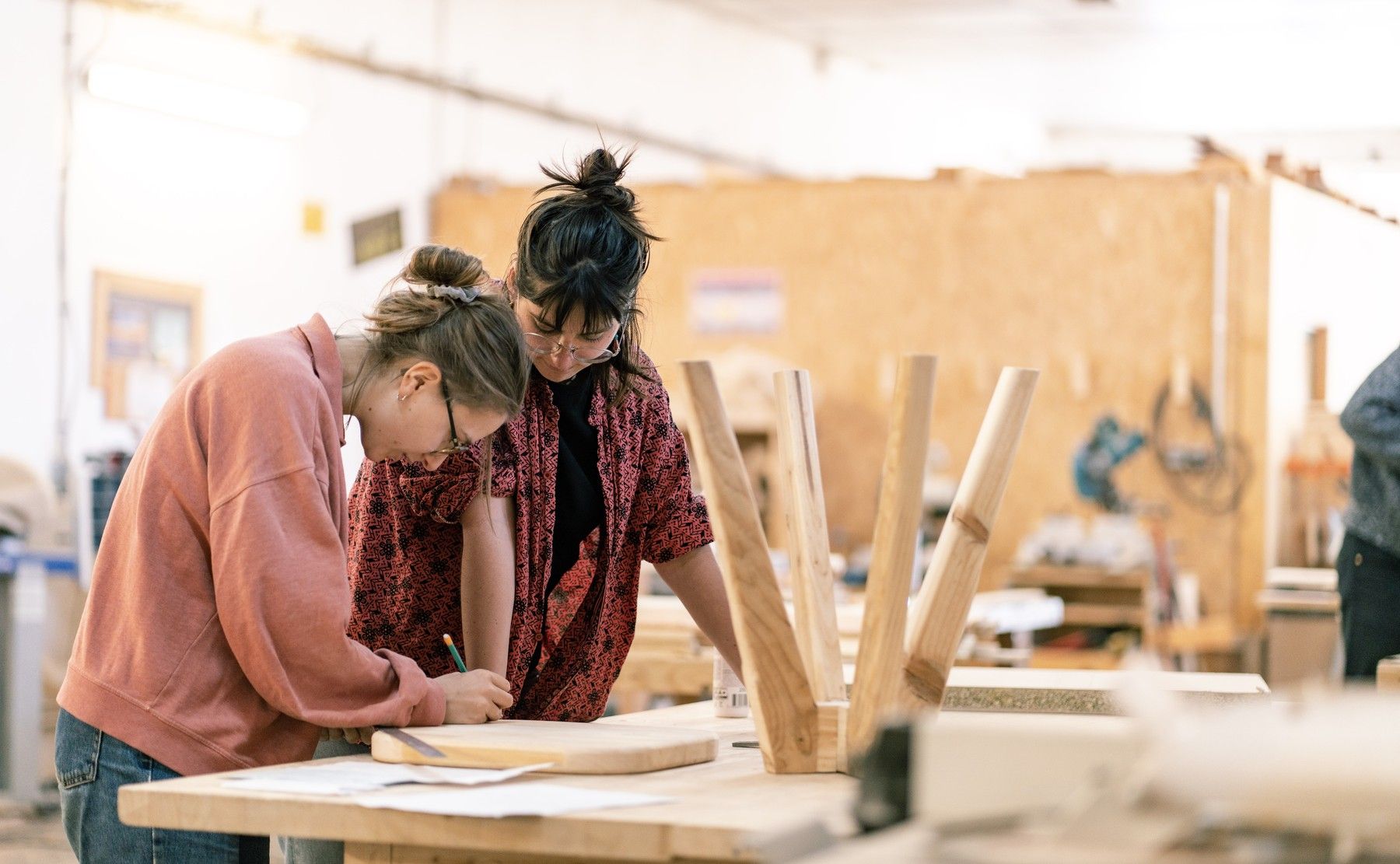 Deux personnes penchées sur un établi de menuiserie tracent au crayon sur une planche ; derrière elles, un tabouret en cours de montage et le mur d'OSB de l'atelier.