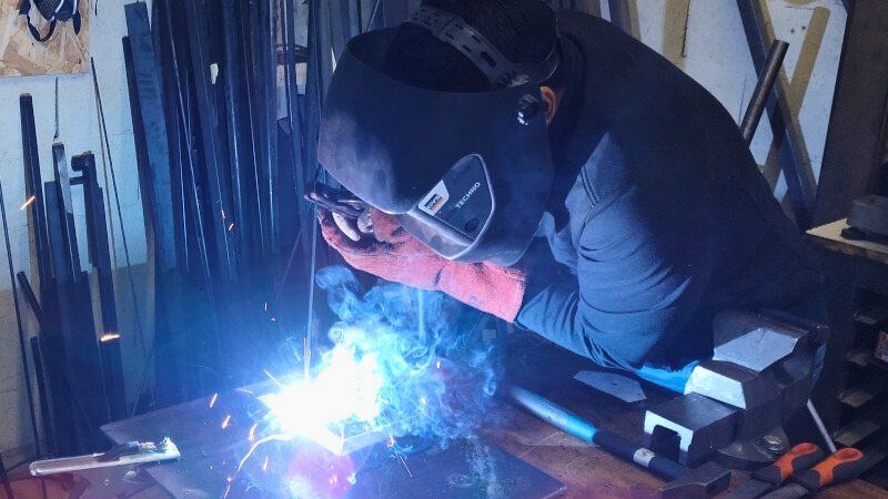 Un soudeur au masque baissé amorce un arc bleu sur une pièce métallique, devant un rack de profilés.