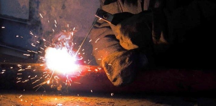 Un soudeur à l'arc, masque baissé et gants de cuir, trace un cordon ; une gerbe d'étincelles éclaire l'établi.