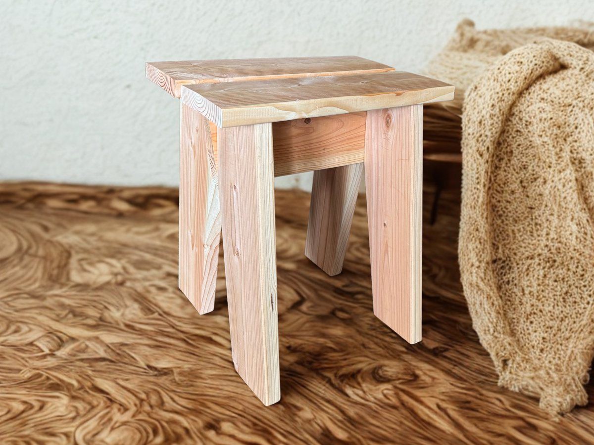 Un tabouret en bois massif clair, assemblé à claire-voie, posé sur un parquet.